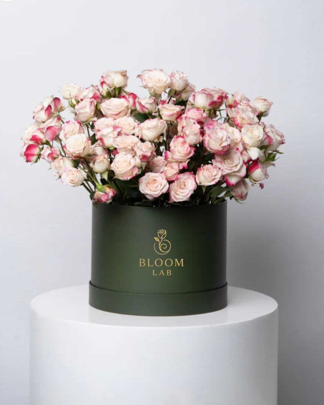 Bouquet of pink spray roses flowers in a green box with 'Bloom Lab' branding on a white pedestal against a light gray background.
